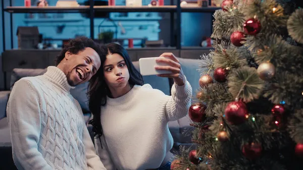 Feliz africano americano hombre tomando selfie con mujer hinchando mejillas cerca de árbol de Navidad - foto de stock