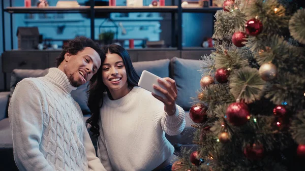 Happy african american woman taking selfie with man sticking out tongue near christmas tree — Stock Photo