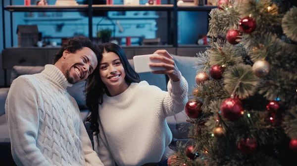 Happy african american man taking selfie with woman near christmas tree — Stock Photo