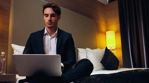 Joven serio hombre de negocios sentado en la cama y el uso de ordenador portátil en la habitación de hotel - foto de stock