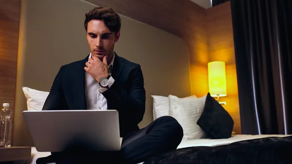 Young pensive businessman sitting on bed and using laptop in hotel room — Stock Photo