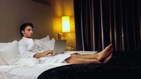 Joven hombre concentrado en albornoz usando portátil en la habitación de hotel - foto de stock
