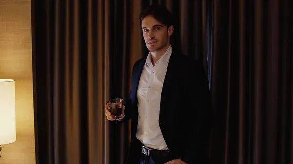 Hombre de negocios guapo sosteniendo un vaso de whisky en la habitación de hotel - foto de stock