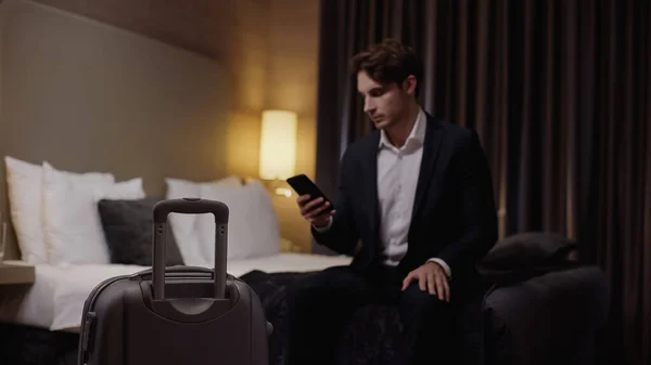 Blurred young businessman holding smartphone near suitcase in hotel room — Stock Photo