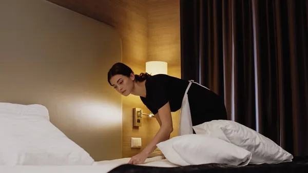 Joven camarera en uniforme y delantal haciendo cama en habitación de hotel - foto de stock