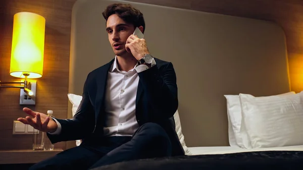 Geschäftsmann im Anzug sitzt auf Bett und gestikuliert, während er im Hotelzimmer mit dem Smartphone spricht — Stockfoto