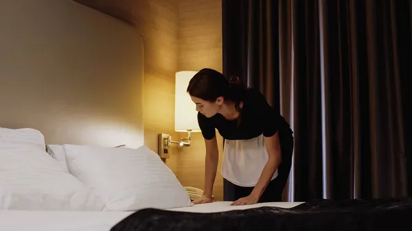 Jovem empregada doméstica em uniforme fazendo cama no quarto de hotel — Fotografia de Stock