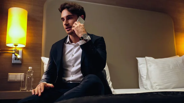 Homem de negócios de terno sentado na cama e falando no smartphone no quarto de hotel — Fotografia de Stock