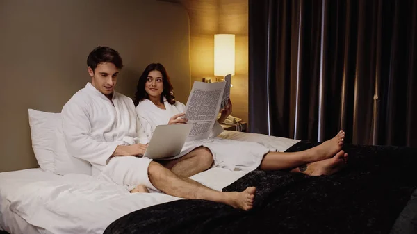 Homem usando laptop perto alegre namorada com jornal descansando no quarto de hotel — Fotografia de Stock