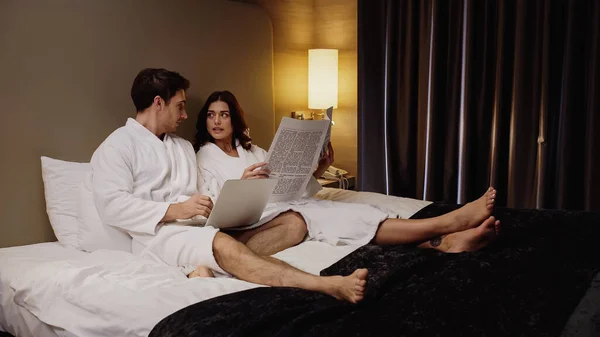 Hombre usando el ordenador portátil y hablando con la novia celebración de periódico en la habitación de hotel - foto de stock