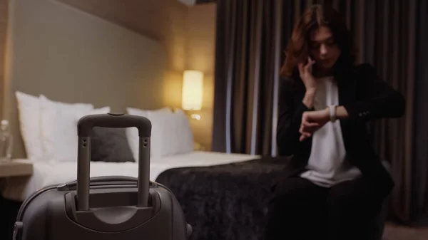 Maleta cerca de la mujer de negocios borrosa hablando en el teléfono inteligente y mirando el reloj de pulsera en la habitación del hotel - foto de stock