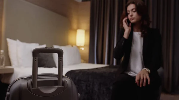 Koffer nahe verschwommener Geschäftsfrau, die im Hotelzimmer mit Smartphone spricht — Stockfoto
