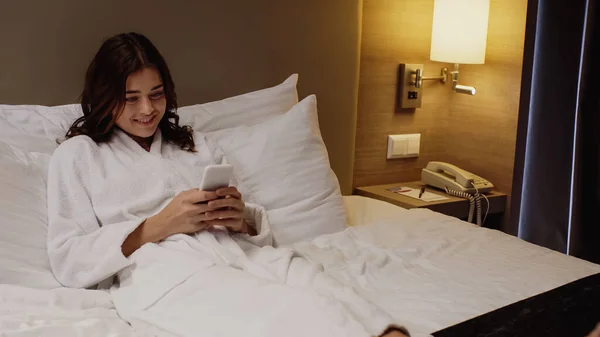 Cheerful woman in bathrobe chatting on smartphone while resting in bed — Stock Photo