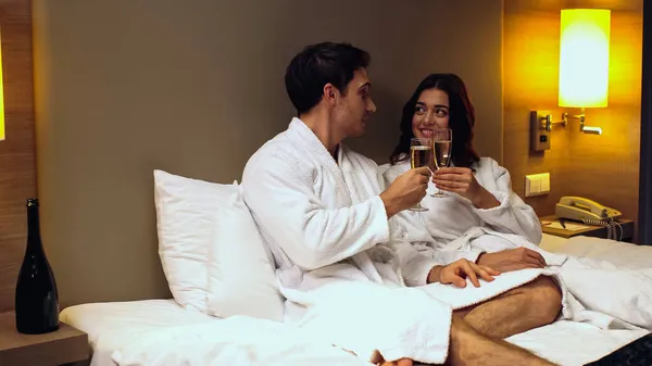 Alegre pareja en albornoces tintineo vasos con champán en habitación de hotel — Stock Photo