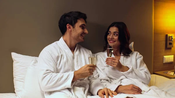 Joyful couple in bathrobes holding glasses with champagne in hotel room — Stock Photo