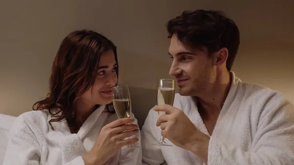 Positive couple in bathrobes holding glasses with champagne in hotel room — Stock Photo