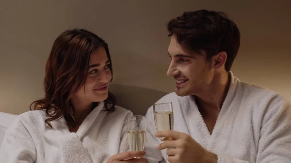 Sonriente pareja joven en albornoces sosteniendo copas con champán en la habitación del hotel - foto de stock