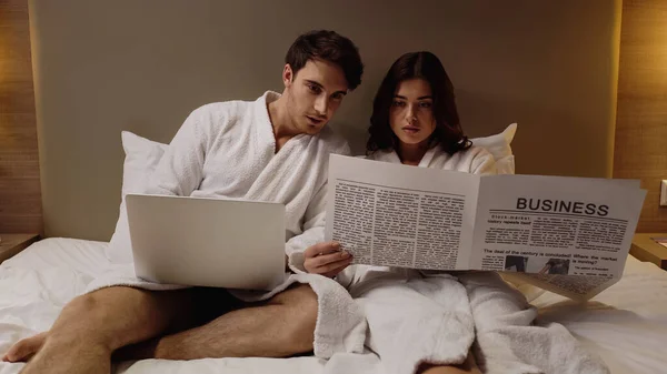 Homem com laptop ler jornal de negócios perto da namorada no quarto de hotel — Fotografia de Stock