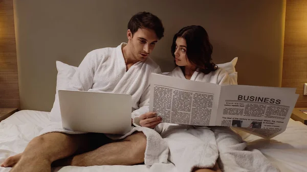 Mann liest mit Laptop Wirtschaftszeitung nahe junger Freundin im Hotelzimmer — Stockfoto
