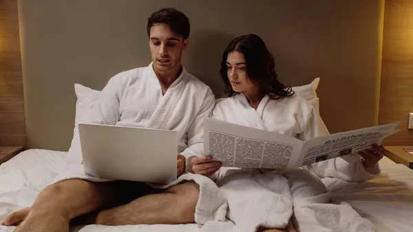 Homme utilisant ordinateur portable près de petite amie avec journal d'affaires reposant dans la chambre d'hôtel — Photo de stock