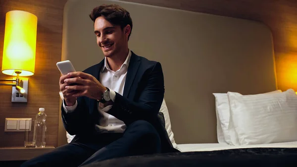 Homem de negócios feliz sentado na cama e conversando no smartphone no quarto de hotel — Fotografia de Stock