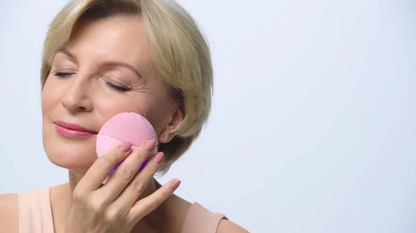 Mujer de mediana edad satisfecho utilizando limpiador de silicona en la cara aislada en blanco — Stock Photo