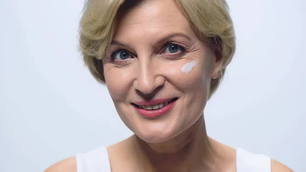 Веселая женщина средних лет с косметическим кремом на лице, смотрящая на камеру, изолированную на белом — стоковое фото