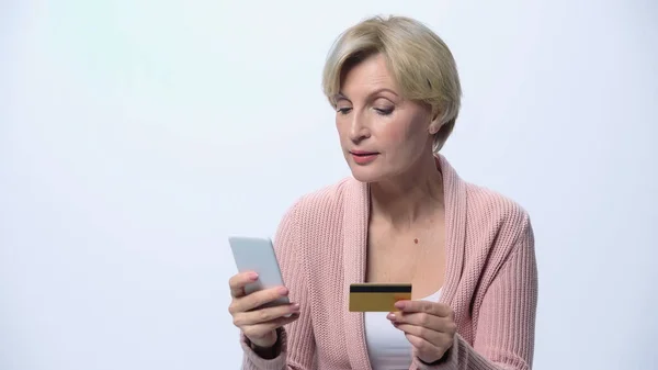 Donna matura che utilizza il telefono cellulare mentre tiene la carta di credito isolata su bianco — Foto stock