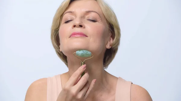 Mature woman with closed eyes massaging face with jade roller isolated on white — Stock Photo