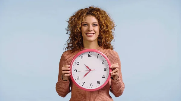 Glückliche Frau mit roten welligen Haaren hält Wanduhr isoliert auf blau — Stockfoto