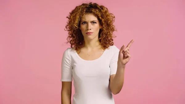 Arrabbiato rossa donna guardando la fotocamera e puntando con il dito isolato su rosa — Foto stock