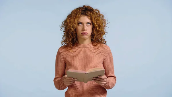 Mujer pelirroja agotado con libro mirando hacia arriba y soplando mejillas aisladas en azul - foto de stock