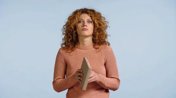 Schockierte junge Frau in rosa Pullover hält Buch in der Hand, während sie isoliert in die Kamera auf blauem Grund blickt — Stockfoto