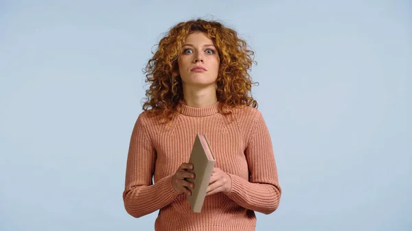 Shocked young woman in pink sweater holding book while looking at camera isolated on blue — Stock Photo