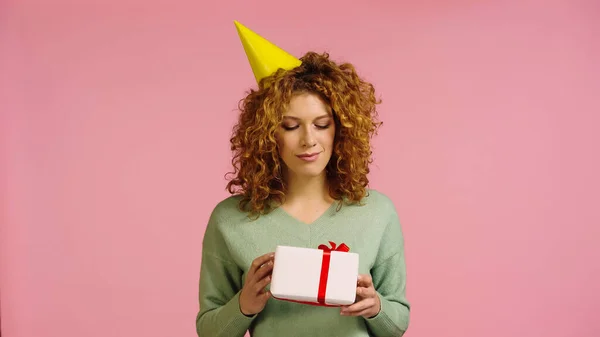 Thoughtful woman in party cap looking at gift box isolated on pink — Stockfoto