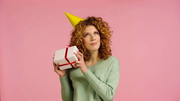 Curious redhead woman in party cap holding gift box isolated on pink — Stockfoto