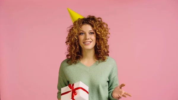Piacere donna in berretto partito guardando la fotocamera mentre tenendo scatola regalo isolato su rosa — Foto stock