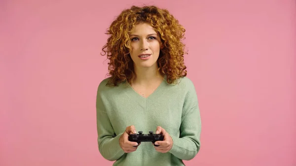 KYIV, UKRAINE - DECEMBER 22, 2021: positive and focused redhead woman playing with gamepad isolated on pink — Foto stock