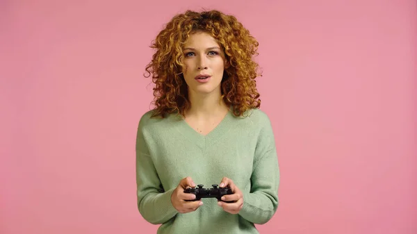 KYIV, UKRAINE - DECEMBER 22, 2021: focused woman with red curly hair playing video game isolated on pink — Foto stock