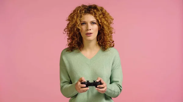 KYIV, UKRAINE - DECEMBER 22, 2021: focused woman with red curly hair playing video game isolated on pink — Stockfoto