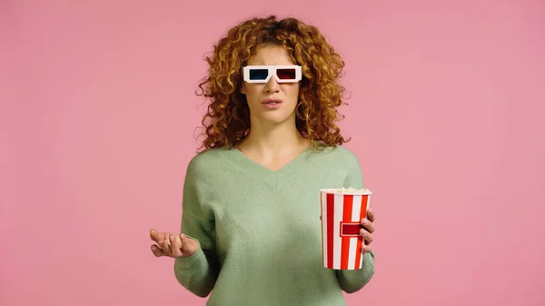 Mujer molesta en gafas 3d sosteniendo palomitas de maíz cubo mientras mira la película aislado en rosa - foto de stock