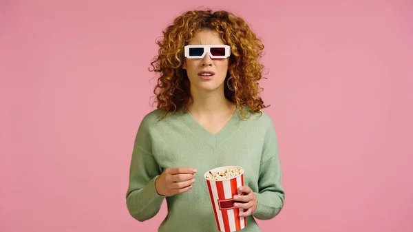 Besorgte Frau in 3D-Gläsern hält Popcorn-Eimer, während sie Film isoliert auf rosa anschaut — Stockfoto