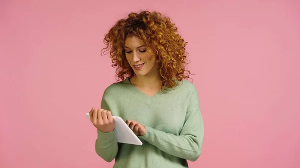 Joven mujer sonriente con el pelo rojo ondulado utilizando tableta digital aislado en rosa - foto de stock