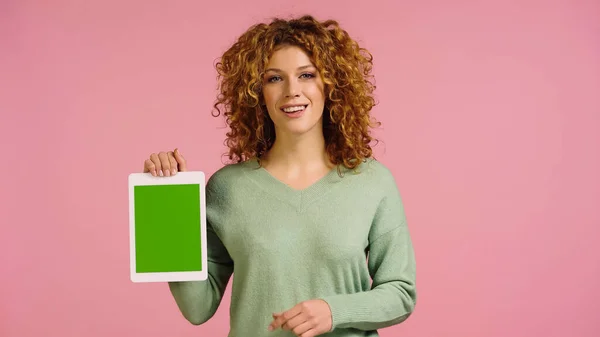 Счастливая рыжая женщина в зеленом свитере проведение цифровой планшет с зеленым экраном изолированы на розовый — стоковое фото
