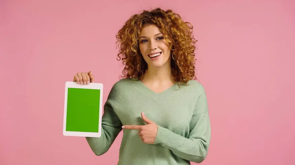 Mujer alegre con el pelo ondulado rojo apuntando a la tableta digital con la pantalla verde aislada en rosa - foto de stock