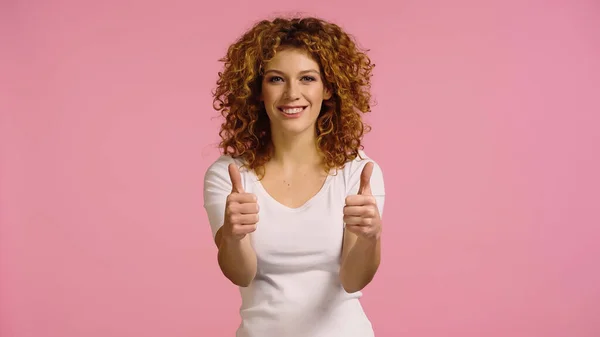 Веселая рыжая женщина в белой футболке показывает большие пальцы вверх, улыбаясь в камеру изолированы на розовый — стоковое фото