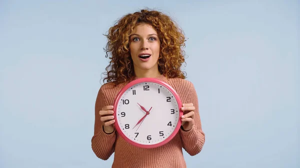 Femme rousse étonnée avec bouche ouverte tenant horloge ronde isolé sur bleu — Photo de stock