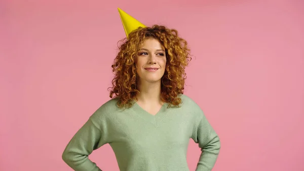 Smiling woman in party cap looking away while standing with hands on hips isolated on pink — Stockfoto