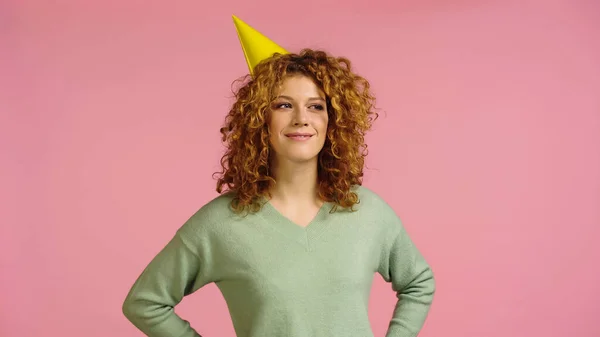 Donna sorridente in berretto partito distogliendo lo sguardo mentre in piedi con le mani sui fianchi isolato su rosa — Foto stock