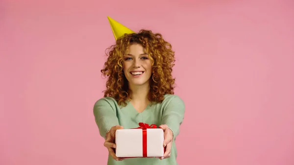Joyful redhead woman in party cap and green jumper showing present isolated on pink — Stockfoto